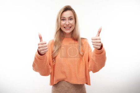 Téléchargez les photos : Heureuse fille blonde montre la main avec les pouces levés et sourit à la caméra isolée sur fond blanc - en image libre de droit