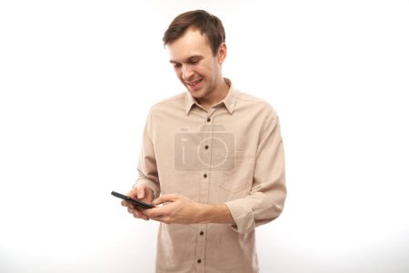 Foto de Retrato de un joven guapo hombre caucásico usando teléfono móvil y sonriendo aislado sobre fondo blanco del estudio, expresión facial alegre - Imagen libre de derechos