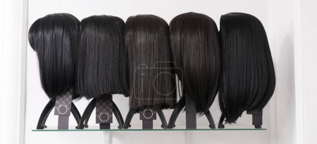 Photo for Showcase of natural looking wigs in different haircut options fixed on the wig holders in beauty salon. Row of mannequin heads with black asian hair on shelf in wig shop - Royalty Free Image