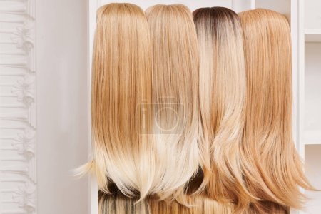 Téléchargez les photos : Vitrine de perruques naturelles dans différentes nuances de blondes fixées sur les porte-perruques dans le salon de beauté. Rangée de têtes de mannequin avec des couleurs de variation cheveux clairs sur l'étagère dans le magasin de perruques - en image libre de droit