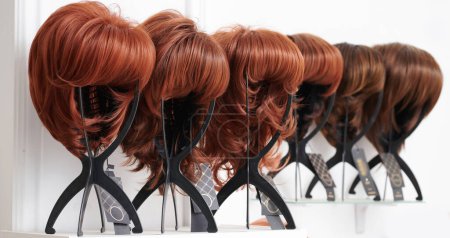 Téléchargez les photos : Vitrine de perruques naturelles dans différentes nuances de brune fixées sur les porte-perruques dans le salon de beauté. Rangée de têtes de mannequin avec variations de couleurs de cheveux sur étagère dans la boutique de perruques - en image libre de droit