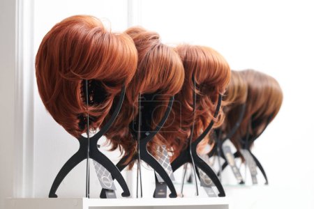 Téléchargez les photos : Vitrine de perruques naturelles dans différentes nuances de brune fixées sur les porte-perruques dans le salon de beauté. Rangée de têtes de mannequin avec variations de couleurs de cheveux sur étagère dans la boutique de perruques - en image libre de droit