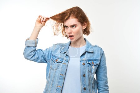 Téléchargez les photos : Portrait rousse jeune femme touchant les cheveux avec le visage mécontent isolé sur fond de studio blanc, concept de cheveux abîmés - en image libre de droit
