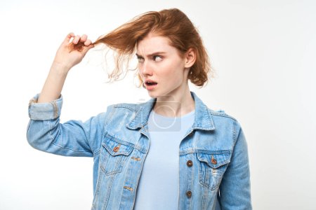 Foto de Retrato pelirroja mujer joven tocando el cabello con la cara descontenta aislado en fondo de estudio blanco, concepto de cabello dañado - Imagen libre de derechos