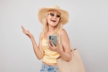 Téléchargez les photos : Portrait femme blonde mature positive en chapeau de paille, haut jaune et lunettes de soleil tenant smartphone et sac à provisions isolés sur fond de studio blanc - en image libre de droit