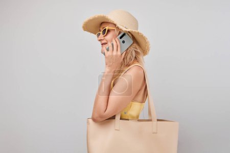 Foto de Retrato de mujer rubia madura positiva en sombrero de paja, parte superior amarilla y gafas de sol que sostienen el teléfono inteligente y la bolsa de compras aislados en el fondo blanco del estudio - Imagen libre de derechos
