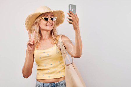 Foto de Retrato de mujer rubia madura positiva en sombrero de paja, parte superior amarilla y gafas de sol que sostienen el teléfono inteligente y la bolsa de compras aislados en el fondo blanco del estudio - Imagen libre de derechos