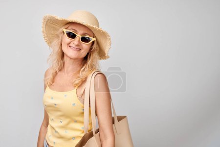 Téléchargez les photos : Portrait d'heureuse belle femme blonde mature en vêtements jaunes d'été, chapeau de paille, lunettes de soleil et sac à provisions souriant isolé sur fond de studio blanc. Concept vacances et voyages - en image libre de droit