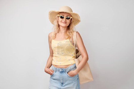 Foto de Retrato de mujer madura rubia hermosa feliz en ropa de verano amarilla, sombrero de paja, gafas de sol y bolsa de compras sonriendo aislado en el fondo del estudio blanco. Concepto de vacaciones y viajes - Imagen libre de derechos