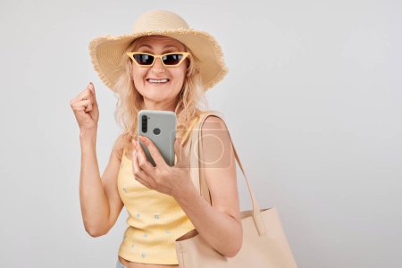 Téléchargez les photos : Portrait femme blonde mature positive en chapeau de paille, haut jaune et lunettes de soleil tenant smartphone et sac à provisions isolés sur fond de studio blanc - en image libre de droit