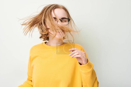 Téléchargez les photos : Portrait de mignonne jeune fille blonde en lunettes et pull jaune secoue la tête et joue avec les cheveux isolés sur fond de studio blanc - en image libre de droit
