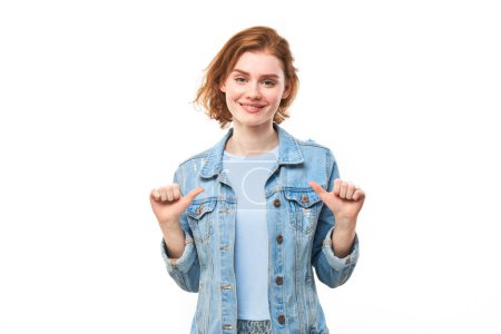Foto de Retrato de una joven pelirroja sonriente, confiada, señalándose con los dedos aislados sobre fondo blanco. Escoge ella misma, concepto de autoestima - Imagen libre de derechos