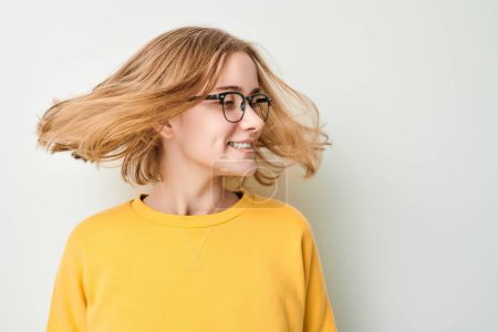 Téléchargez les photos : Portrait de mignonne jeune fille blonde en lunettes et pull jaune secoue la tête et joue avec les cheveux isolés sur fond de studio blanc - en image libre de droit