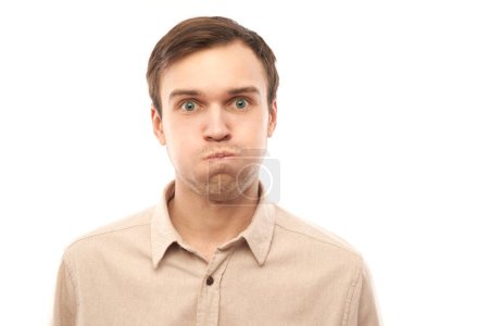 Téléchargez les photos : Portrait de jeune homme caucasien gonflant les joues avec un drôle de visage isolé sur fond de studio blanc. Concept d'humour, bouche gonflée avec l'expression folle d'air - en image libre de droit