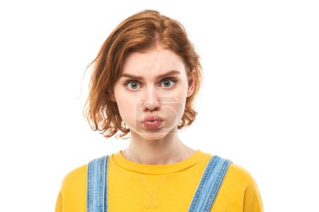 Téléchargez les photos : Portrait de jeune femme rousse gonflant les joues avec un drôle de visage isolé sur fond de studio blanc. Concept d'humour, bouche gonflée avec l'expression folle d'air - en image libre de droit