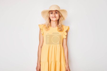 Foto de Retrato de mujer madura rubia hermosa feliz en vestido de verano amarillo y sombrero de paja sonriendo alegremente aislado sobre fondo de estudio blanco. Concepto de vacaciones y viajes - Imagen libre de derechos