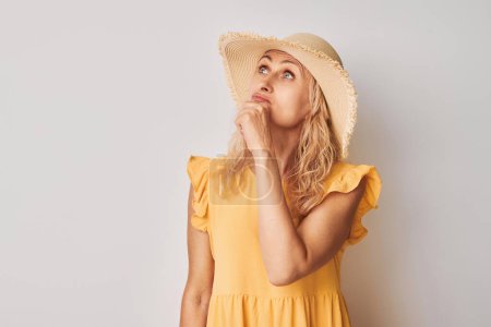 Foto de Retrato de mujer madura inteligente en ropa amarilla de verano tocando la barbilla pensando elige dudas aisladas en el fondo del estudio blanco con spac copia - Imagen libre de derechos