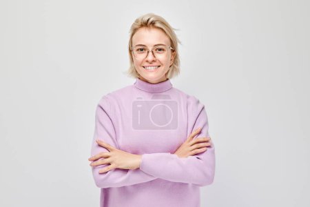 Foto de Retrato de mujer rubia joven caucásica sonriendo alegremente aislada sobre fondo blanco del estudio. Chica feliz en suéter lila y gafas con expresión de cara alegre - Imagen libre de derechos