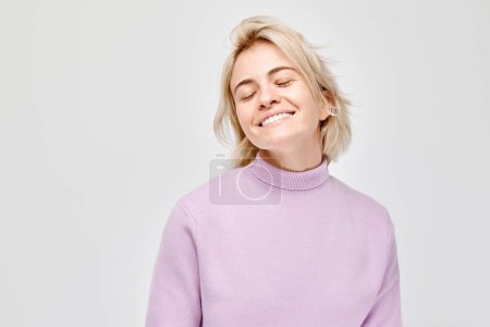 Téléchargez les photos : Portrait Femme blanche jeune blonde souriante joyeusement isolée sur fond de studio blanc. Fille heureuse en pull lilas avec expressio visage heureux - en image libre de droit