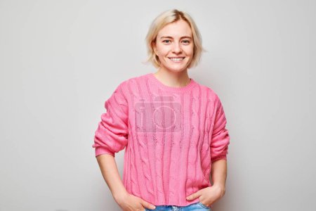 Téléchargez les photos : Portrait Femme blanche jeune blonde souriante joyeusement isolée sur fond de studio blanc. Fille heureuse en pull rose avec expression de visage heureux - en image libre de droit