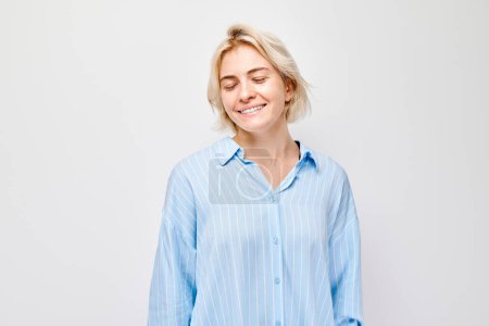Foto de Retrato de mujer rubia joven caucásica sonriendo alegremente aislada sobre fondo blanco del estudio. Chica feliz en camisa azul con expresión de cara alegre - Imagen libre de derechos