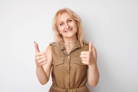 Photo for Portrait of mature blond woman smiling joyfully showing thumbs up gesture isolated on white studio background. Approves good choice, right decision - Royalty Free Image