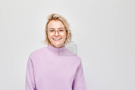 Téléchargez les photos : Portrait Femme blanche jeune blonde souriante joyeusement isolée sur fond de studio blanc. Fille heureuse en pull lilas et lunettes avec expression joyeuse du visage - en image libre de droit