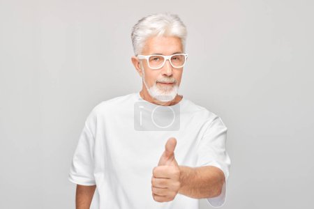 Photo for Portrait of mature man in white T-shirt and glasses smiling joyfully showing thumbs up gesture isolated on white studio background. Approves good choice, right decisio - Royalty Free Image