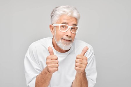 Photo for Portrait of mature man in white T-shirt and glasses smiling joyfully showing thumbs up gesture isolated on white studio background. Approves good choice, right decisio - Royalty Free Image
