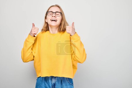 Retrato de una joven estudiante sonriendo alegremente mostrando un gesto de pulgares hacia arriba aislado sobre fondo blanco del estudio. Aprueba la buena elección, la decisión correcta