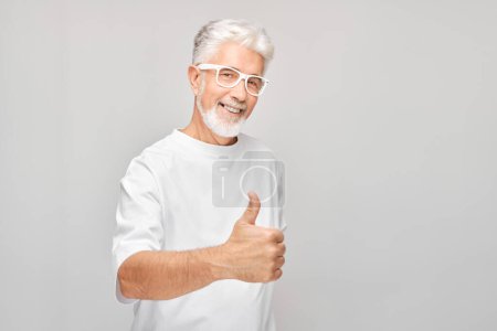 Foto de Retrato de hombre maduro en camiseta blanca y gafas sonriendo alegremente mostrando los pulgares hacia arriba gesto aislado sobre fondo blanco del estudio. Aprueba la buena elección, la decisión correcta - Imagen libre de derechos