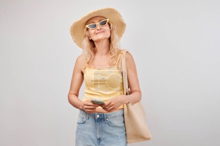 Photo for Portrait positive mature blond woman in straw hat, yellow top and sunglasses holding smartphone and shopping bag isolated on white studio background - Royalty Free Image