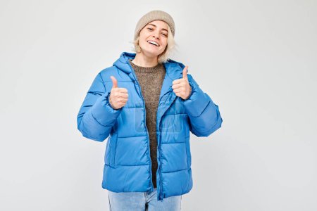 Foto de Retrato de una joven rubia en traje de invierno sonriendo alegremente mostrando el gesto de los pulgares hacia arriba aislado en el fondo blanco del estudio. Aprueba la buena elección, la decisión correcta - Imagen libre de derechos