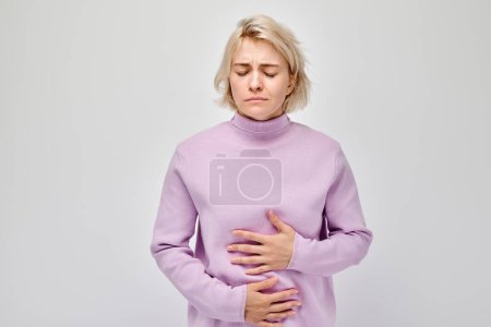 Portrait of young blonde woman suffering from stomach pain isolated on white studio background. Intestinal and stomach problems concep