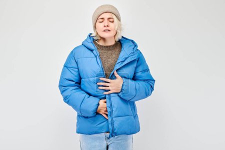 Téléchargez les photos : Portrait de jeune femme en veste d'hiver et chapeau souffrant de douleurs à l'estomac isolé sur fond de studio blanc. Problèmes intestinaux et gastriques concéder - en image libre de droit