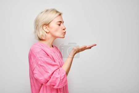 Téléchargez les photos : Portrait de jeune femme blonde en pull rose envoyant un baiser d'air, tenant quelque chose dans la paume, démontrant un produit isolé sur fond de studio blanc - en image libre de droit