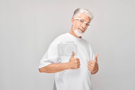 Foto de Retrato de hombre maduro en camiseta blanca y gafas sonriendo alegremente mostrando los pulgares hacia arriba gesto aislado sobre fondo blanco del estudio. Aprueba la buena elección, la decisión correcta - Imagen libre de derechos