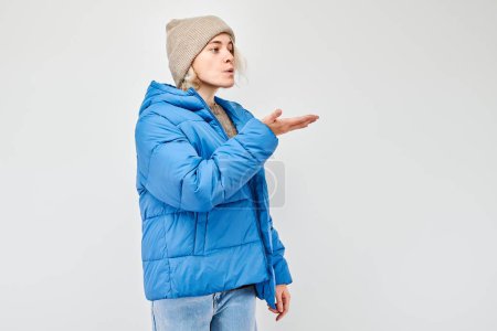 Photo for Portrait of young blond woman in winter jacket sending air kiss, holding something in palm, demonstrating product isolated on white studio background - Royalty Free Image