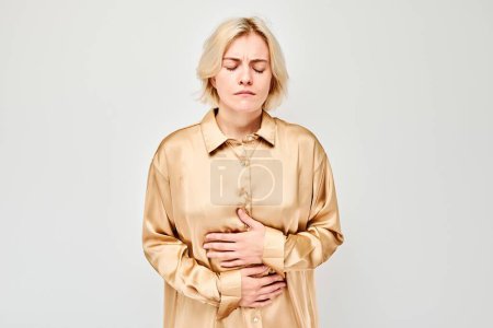 Téléchargez les photos : Portrait de jeune femme blonde souffrant de douleurs abdominales isolées sur fond de studio blanc. Problèmes intestinaux et gastriques concéder - en image libre de droit