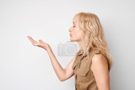 Téléchargez les photos : Portrait de femme mature blonde envoyant un baiser d'air, tenant quelque chose dans la paume, démontrant un produit isolé sur fond de studio blanc - en image libre de droit
