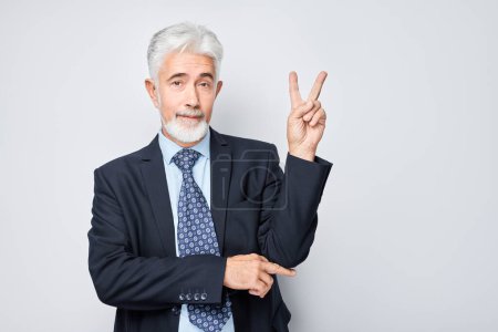 Téléchargez les photos : Portrait d'un homme d'affaires adulte positif en costume montrant un geste de paix et d'amour avec les doigts isolés sur fond de studio blanc. Selfie pose, bonjour mon ami concéder - en image libre de droit