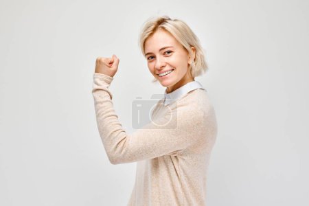 Foto de Retrato de una joven rubia confiada mostrando bíceps, demostrando fuerza aislada sobre fondo blanco del estudio. Afortunado ganador cóncavo - Imagen libre de derechos