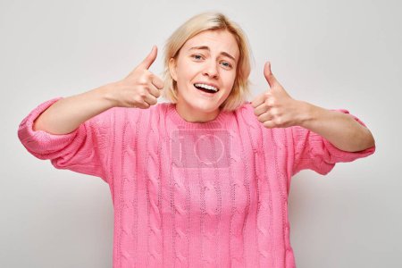Foto de Retrato de una joven rubia en suéter rosa sonriendo alegremente mostrando el gesto de los pulgares hacia arriba aislado en el fondo blanco del estudio. Aprueba la buena elección, la decisión correcta - Imagen libre de derechos