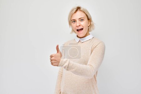 Téléchargez les photos : Portrait de jeune femme blonde en tenue décontractée souriant montrant joyeusement les pouces vers le haut geste isolé sur fond de studio blanc. Approuve le bon choix, la bonne décision - en image libre de droit