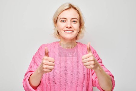 Téléchargez les photos : Portrait de jeune femme blonde en pull rose souriant montrant joyeusement les pouces vers le haut geste isolé sur fond de studio blanc. Approuve le bon choix, la bonne décision - en image libre de droit