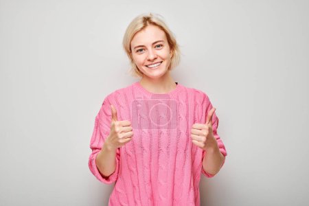 Photo for Portrait of young blond woman in pink sweater smiling joyfully showing thumbs up gesture isolated on white studio background. Approves good choice, right decision - Royalty Free Image