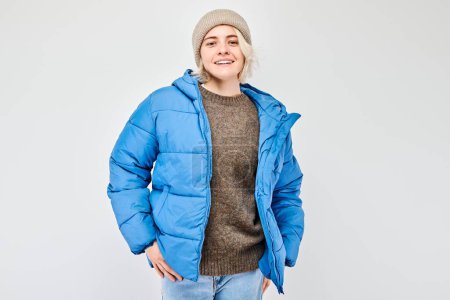 Photo for Portrait Caucasian young blond woman smiling joyfully isolated on white studio background. Happy girl in winter blue jacket and hat with glad face expression - Royalty Free Image