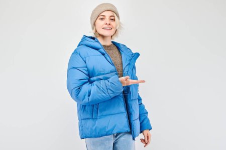 Foto de Mujer joven con chaqueta azul de invierno sosteniendo algo en la mano, demostrando espacio vacío para el producto o el texto aislado en el fondo del estudio blanco - Imagen libre de derechos