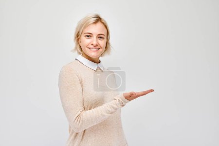 Foto de Mujer rubia joven en casual sosteniendo algo en la mano, demostrando espacio vacío para producto o texto aislado en fondo de estudio blanco - Imagen libre de derechos