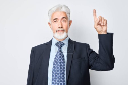 Téléchargez les photos : Homme mûr aux cheveux gris 60 ans en costume d'affaires pointant le doigt vers le haut, démontrant un espace vide pour le produit ou le texte isolé sur fond de studio blanc - en image libre de droit